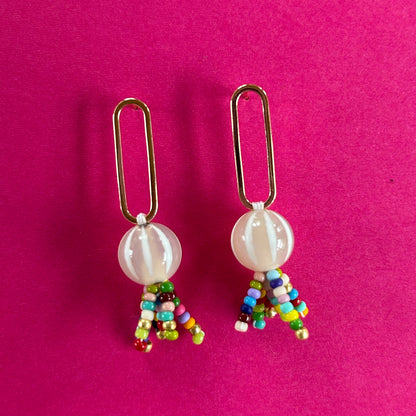 A pair of handmade paperclip earrings with gold-tone metal paperclips, white marbled beads, and dangling multicolor seed bead tassels, displayed on a vibrant pink background.