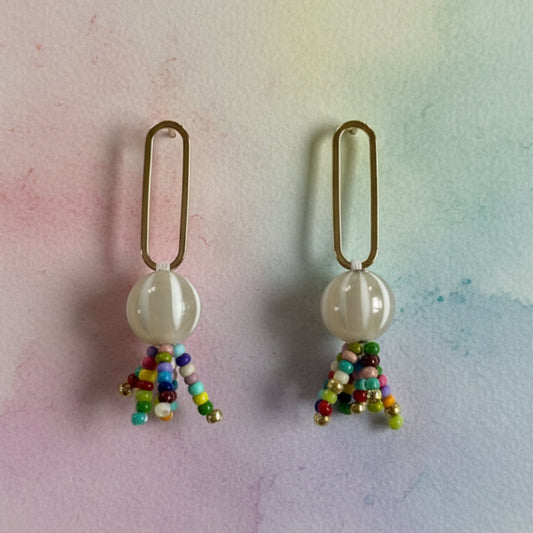 A pair of handmade dangle earrings featuring a small gold-colored loop, a round off-white bead, and a dangling tassel made of tiny multicolored beads, set against a plain light gray background.