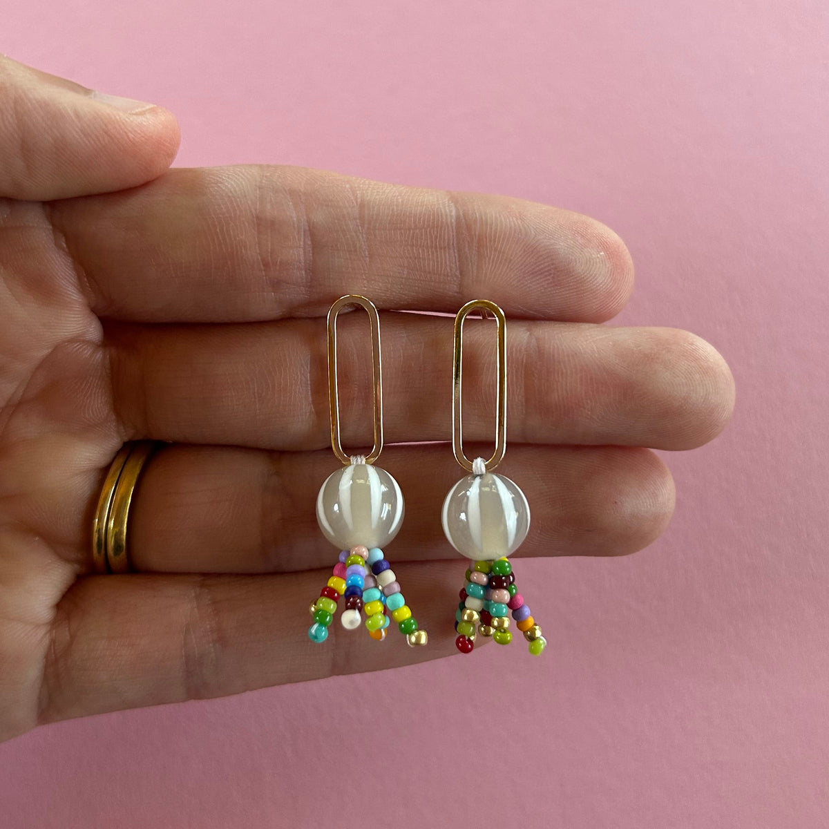 A pair of gold-tone dangle earrings with round white beads and small multicolored beaded fringe, held between a person's fingers against a solid pink background.