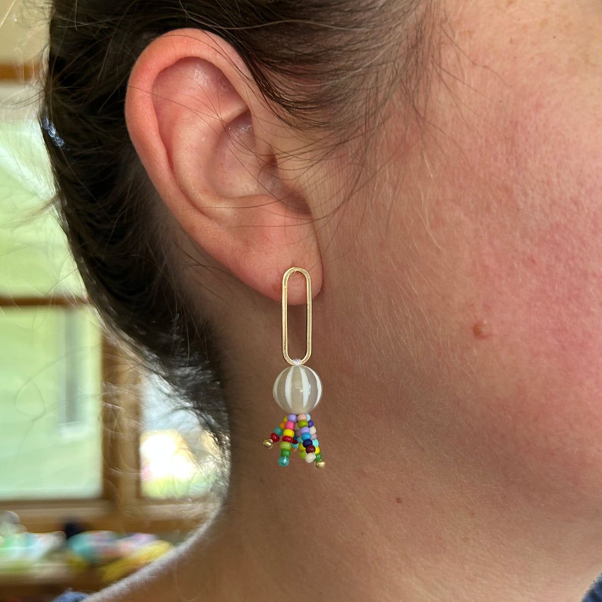 A close-up side profile of a woman wearing a gold paperclip-shaped dangle earring with a clear round bead and small multicolored beads at the bottom.