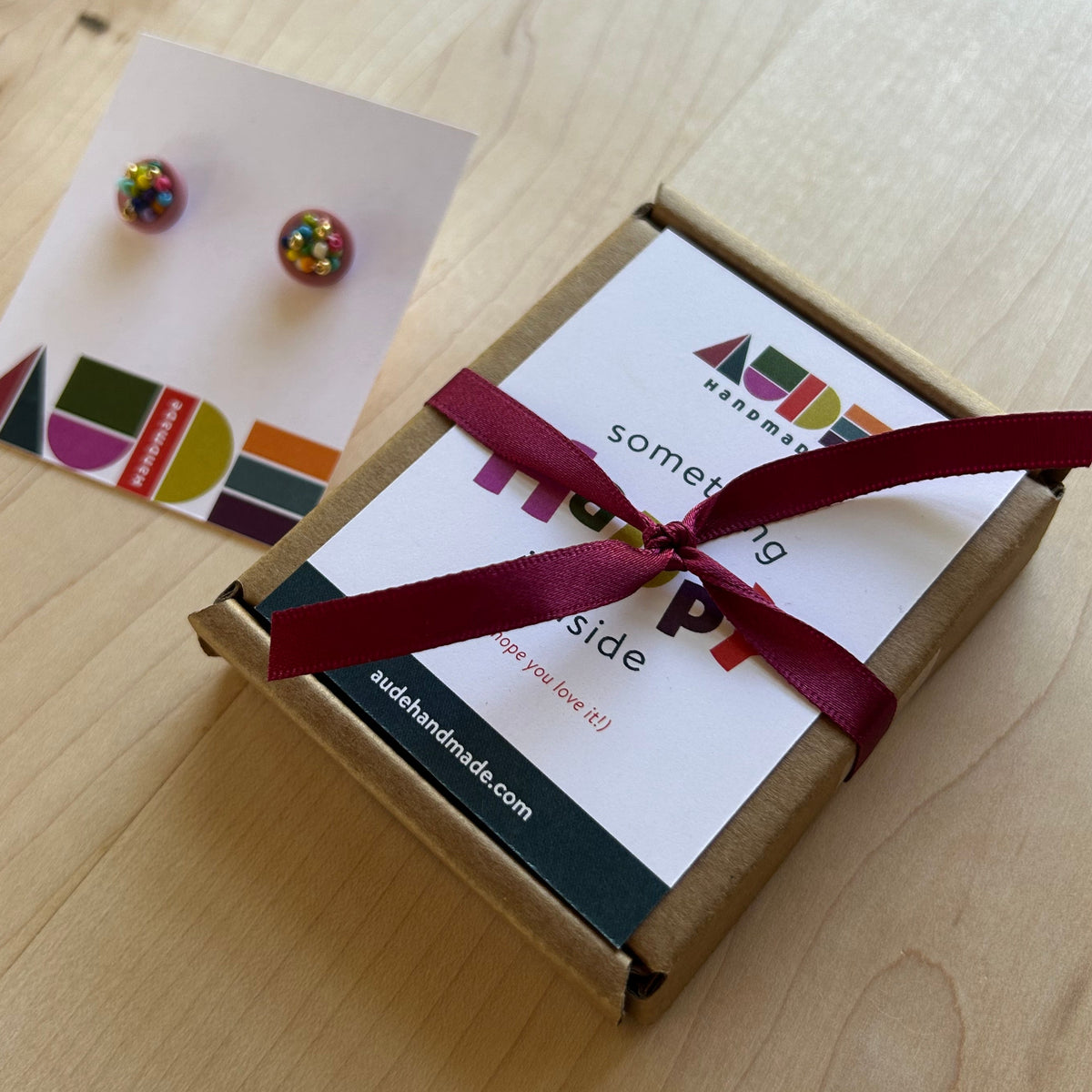 A small kraft paper gift box tied with a burgundy ribbon sits on a light wood table next to a white card holding two round, multicolored stud earrings. The box lid has a colorful logo and text, and the card has geometric color blocks.
