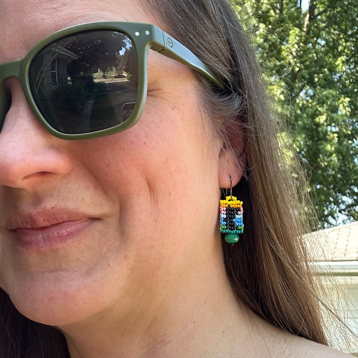 A close-up of a woman wearing olive green sunglasses and a colorful beaded drop earring outdoors with trees and a house in the background.