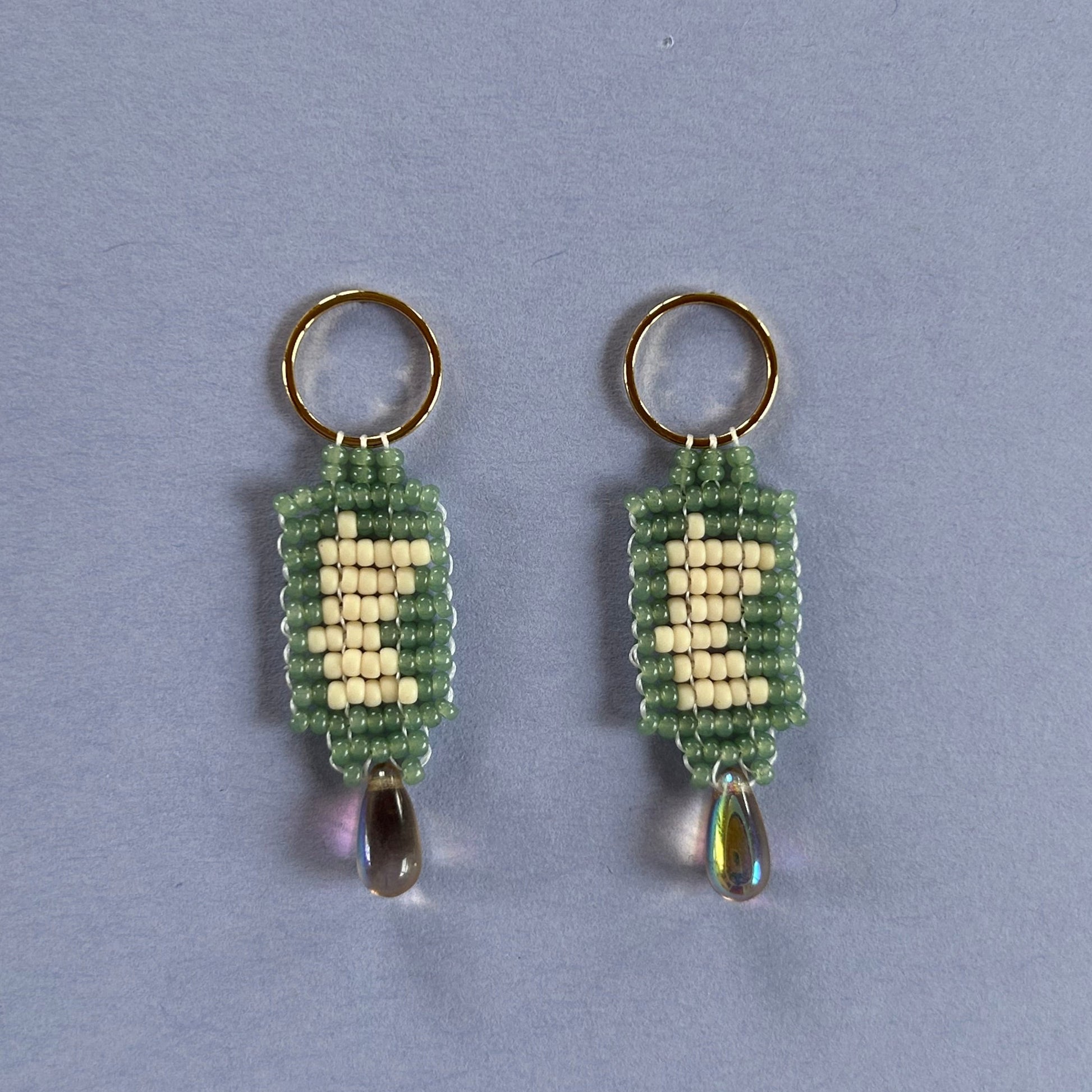 A pair of handmade beaded earrings featuring a green and cream woven pattern, with a small iridescent teardrop bead hanging from each ring, set against a solid light blue background.