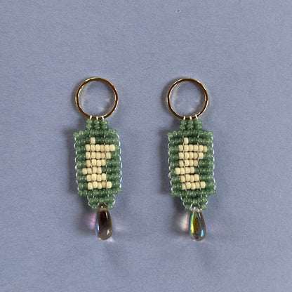 A pair of handmade beaded earrings featuring a green and cream woven pattern, with a small iridescent teardrop bead hanging from each ring, set against a solid light blue background.