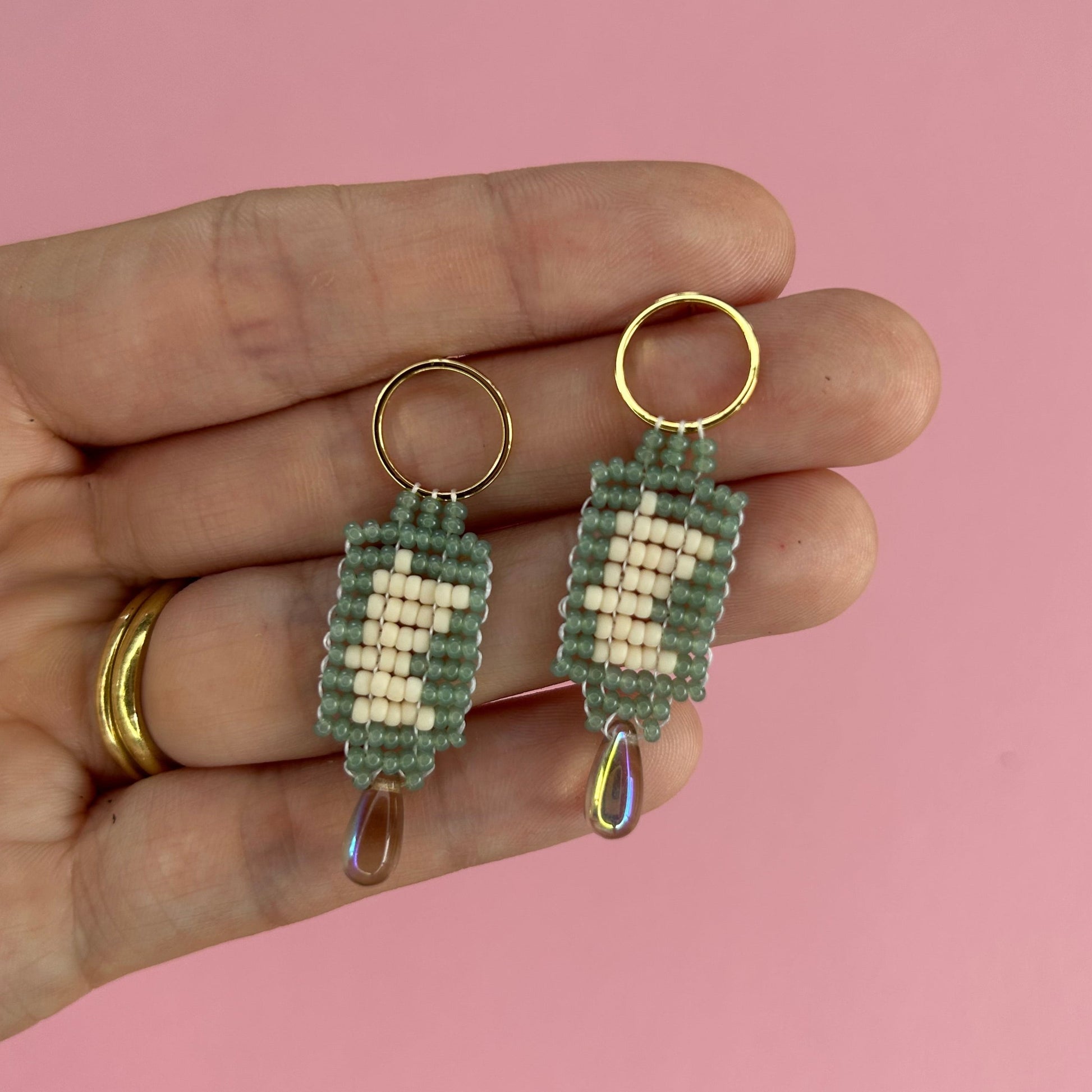 A hand holding a pair of handmade beaded earrings against a solid pink background. The earrings feature sage green and cream woven rectangular beads with gold-tone posts and dangling iridescent teardrop charms.