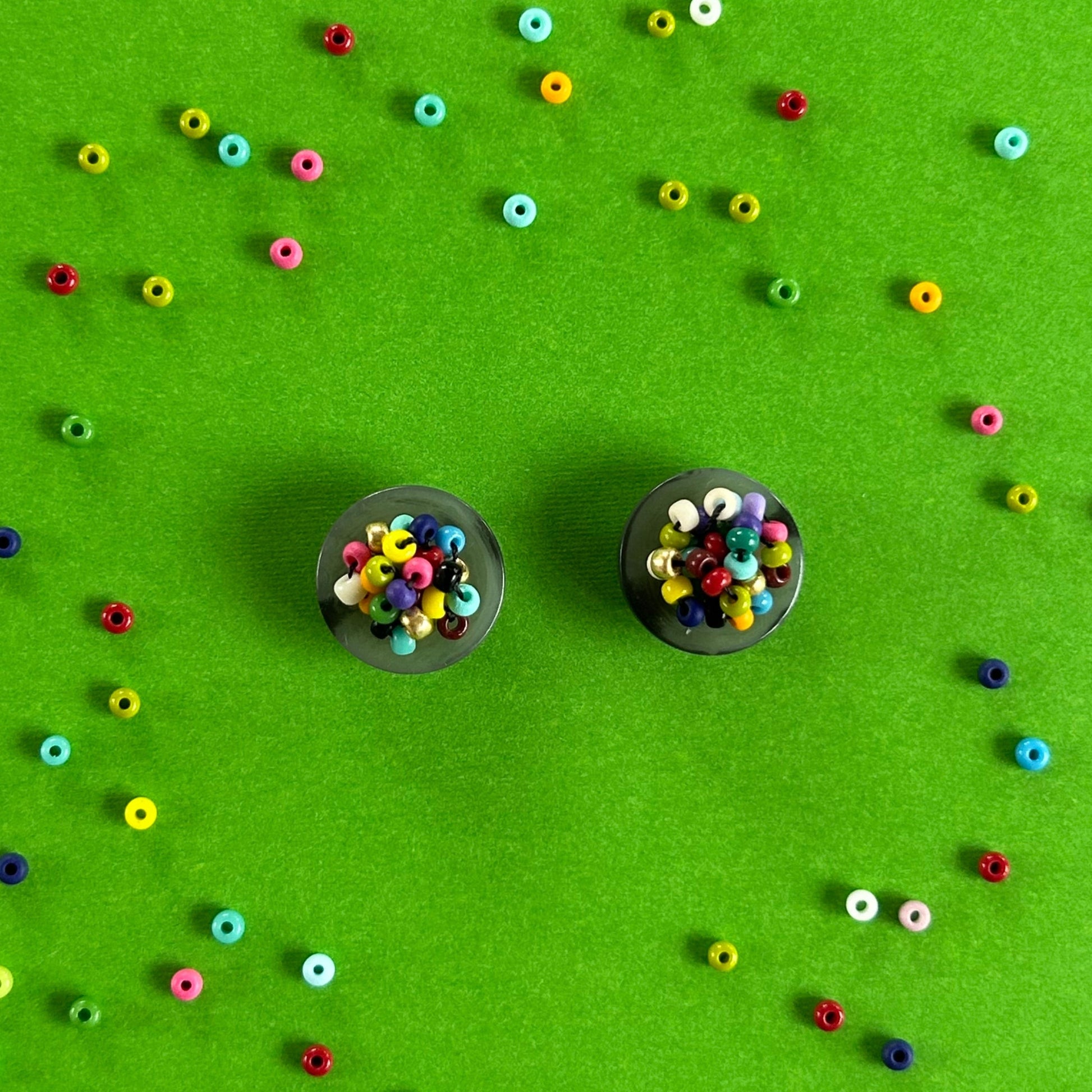 Two small black circular earrings filled with multicolored seed beads, arranged on a bright green background with scattered loose beads forming a decorative pattern around them.