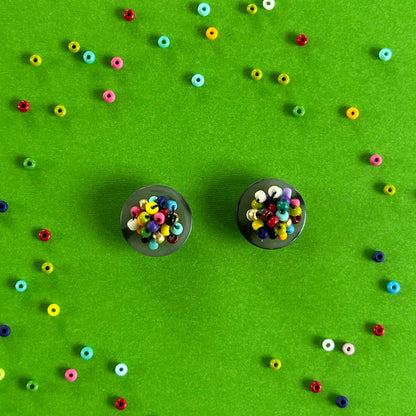 Two small black circular earrings filled with multicolored seed beads, arranged on a bright green background with scattered loose beads forming a decorative pattern around them.