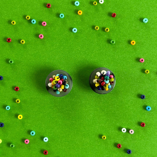 Two small black circular earrings filled with multicolored seed beads, arranged on a bright green background with scattered loose beads forming a decorative pattern around them.