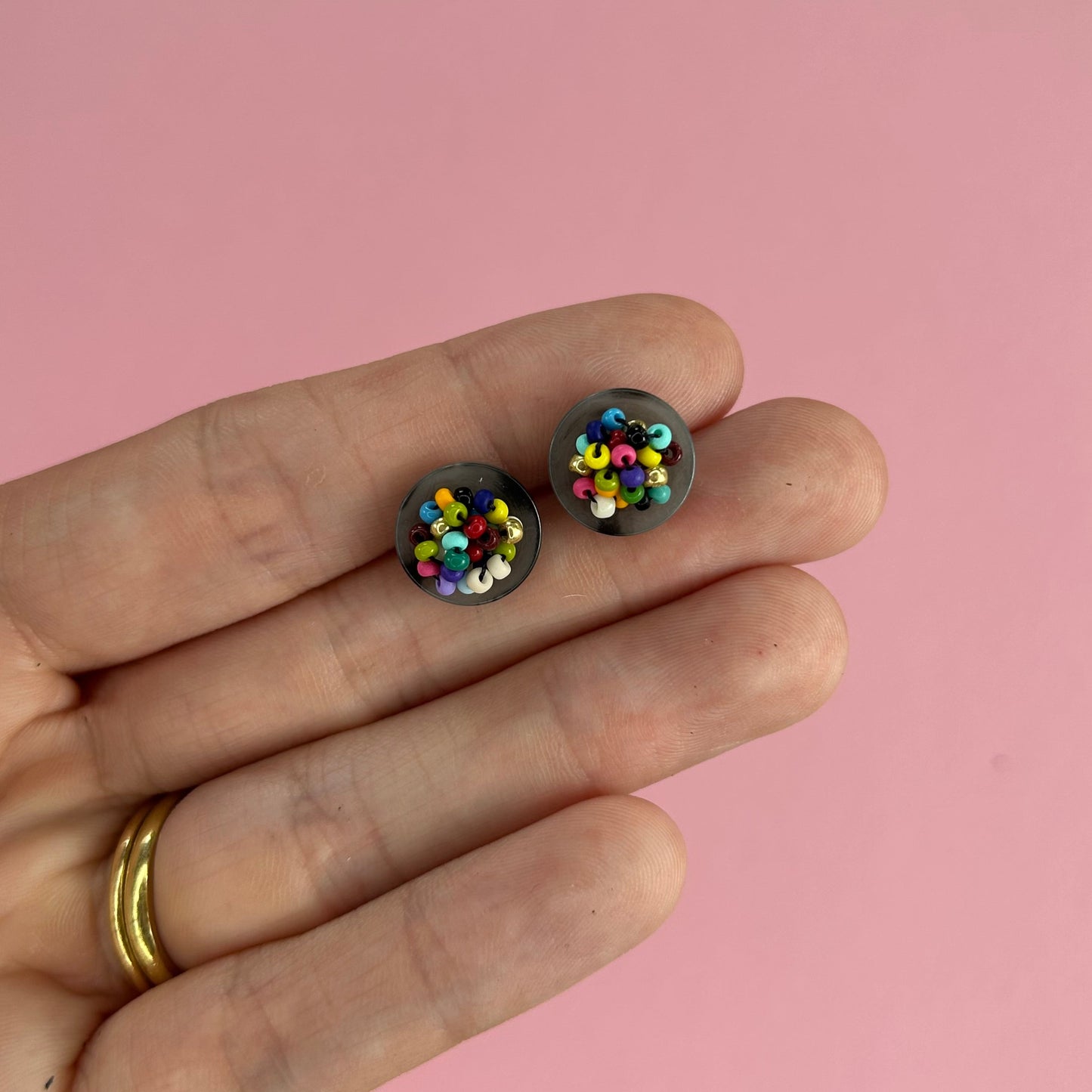 A close-up of a hand holding two small round stud earrings filled with colorful beads, set against a solid pink background.