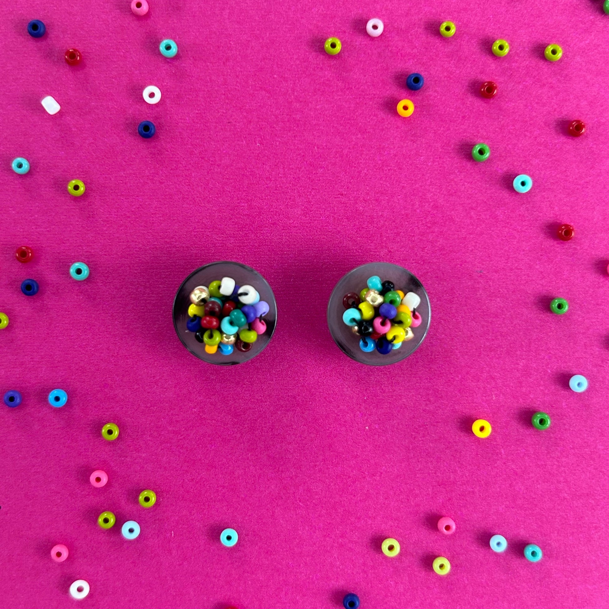 Two round black buttons filled with colorful seed beads, arranged on a bright pink background scattered with loose matching beads.