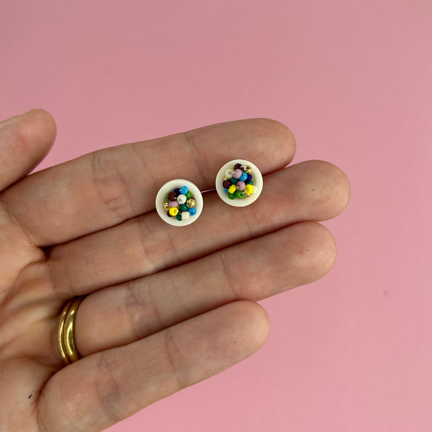 A close-up of a hand holding a pair of tiny white stud earrings filled with small multicolored beads, set against a solid pink background.