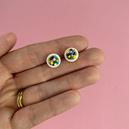 A close-up of a hand holding a pair of tiny white stud earrings filled with small multicolored beads, set against a solid pink background.