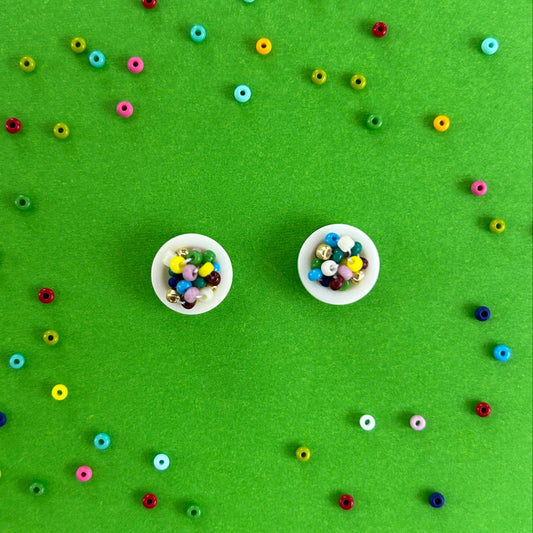 Two small white earrings filled with colorful round craft beads, arranged on a bright green background with scattered loose beads around them.
