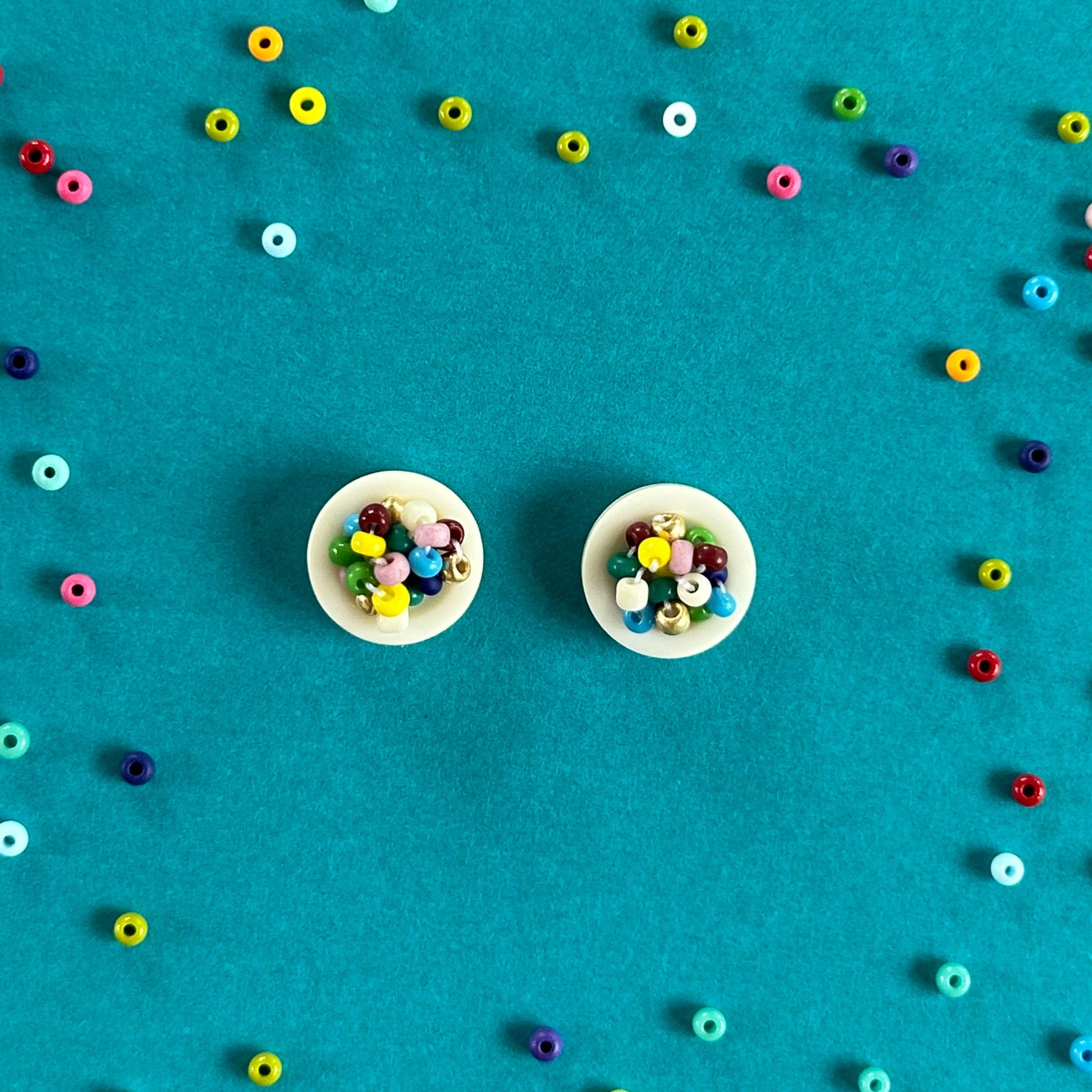 Two small round white buttons filled with colorful loose beads, arranged on a bright turquoise background with scattered loose beads forming a loose circle around them.