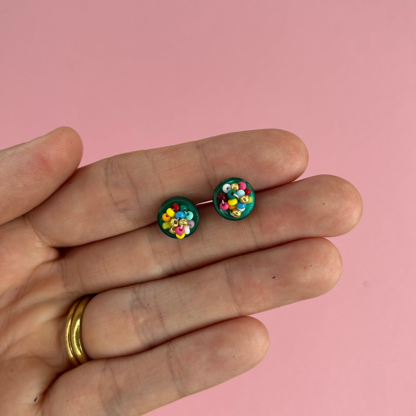 A close-up of a hand holding a pair of small round stud earrings with colorful confetti-like details on a solid pink background.