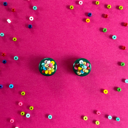 Two round black earrings filled with colorful seed beads, arranged on a bright pink background scattered with loose multicolored beads.