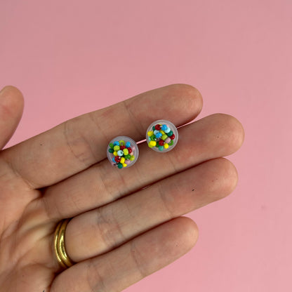A hand holding a pair of stud earrings with colorful confetti-like details inside clear domed settings, set against a solid pink background.