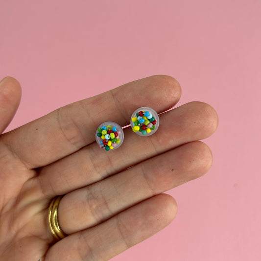 A hand holding a pair of stud earrings with colorful confetti-like details inside clear domed settings, set against a solid pink background.