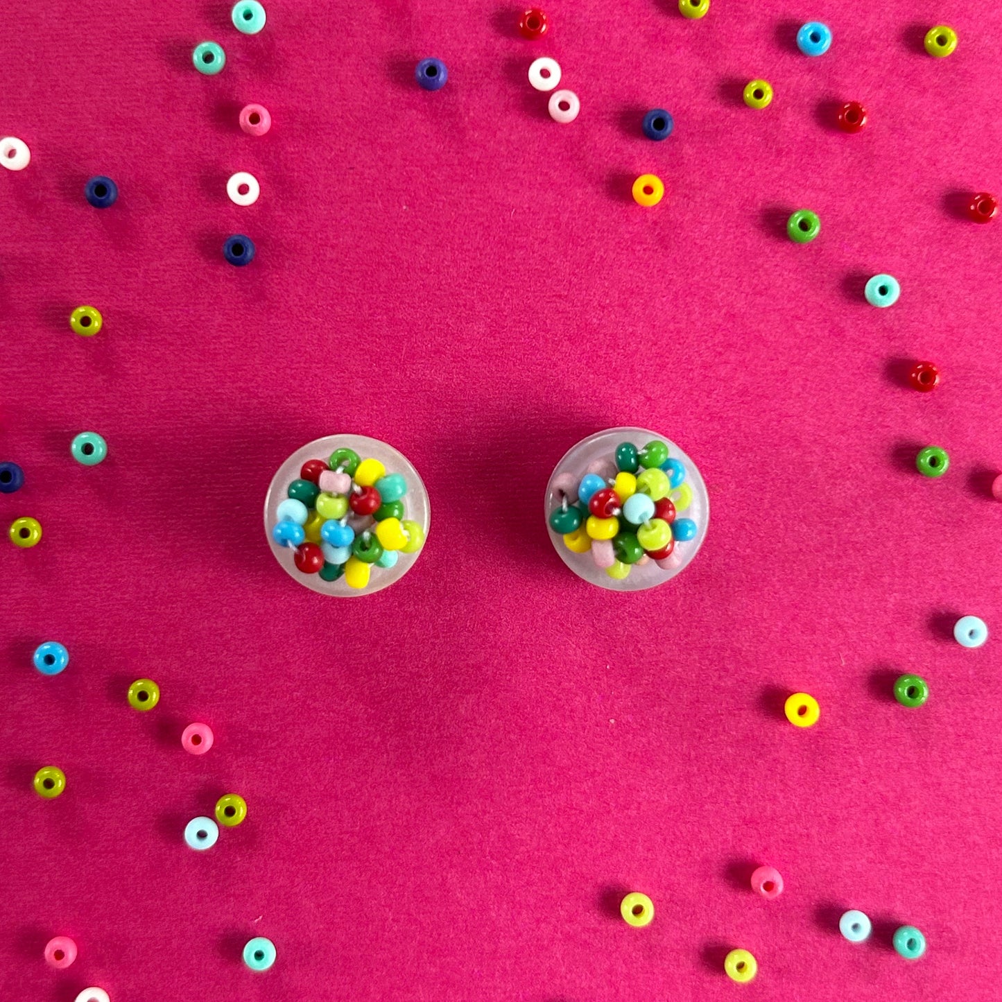 Two small clear plastic cups filled with colorful round seed beads, arranged on a bright pink background with scattered loose beads around them.