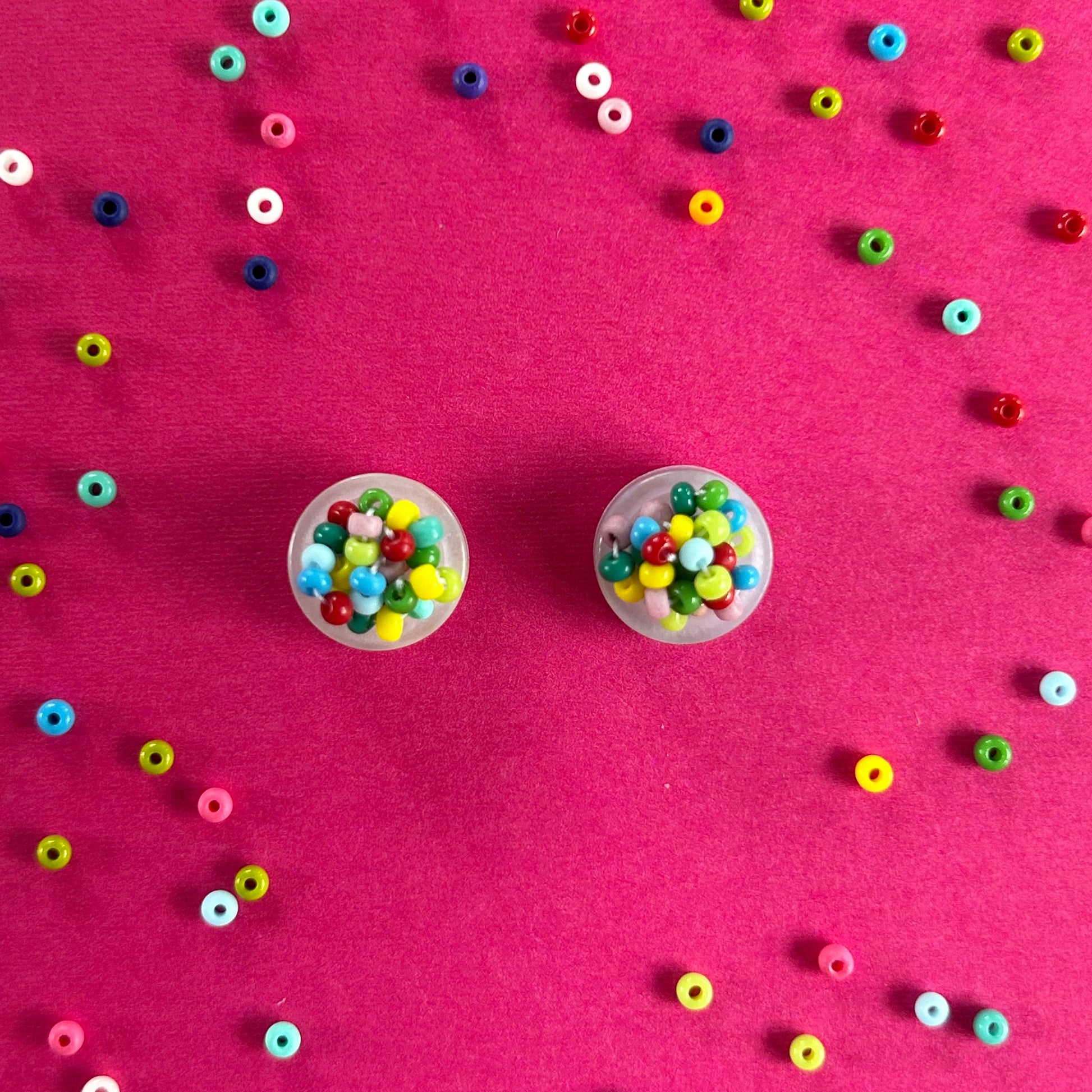 Two small clear plastic cups filled with colorful round seed beads, arranged on a bright pink background with scattered loose beads around them.