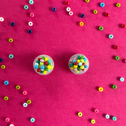 Two small clear plastic cups filled with colorful round seed beads, arranged on a bright pink background with scattered loose beads around them.