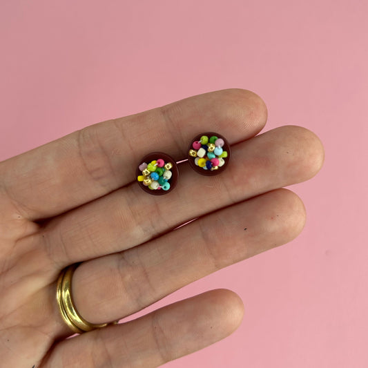 Hand holding two stud earrings with colorful seed beads on a light pink background.