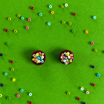 Two small round brown buttons filled with colorful seed beads, arranged on a bright green background with scattered loose beads forming a loose circle around them.