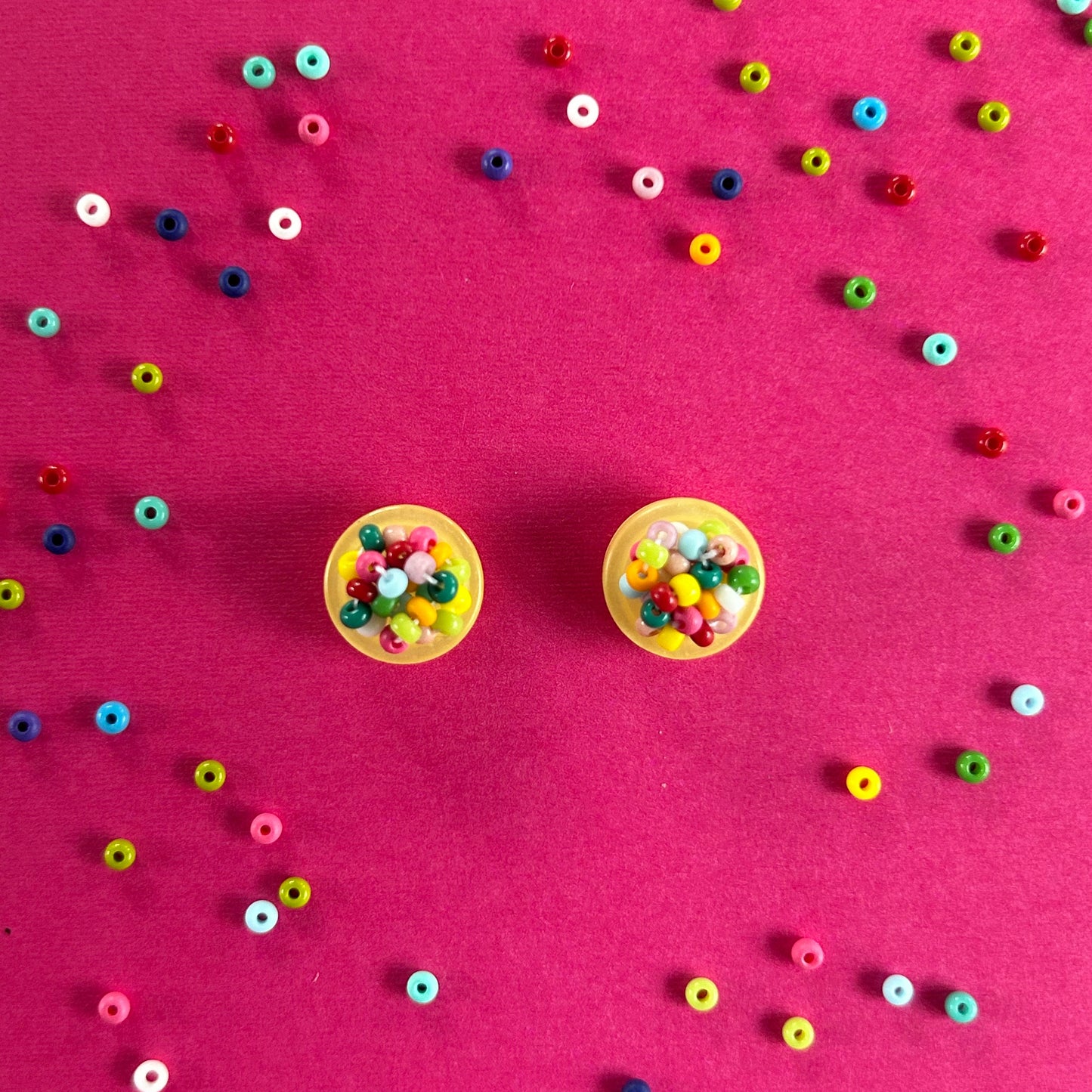 Two small yellow circular earrings filled with colorful round beads, arranged on a bright pink background with scattered loose multicolored beads around them.