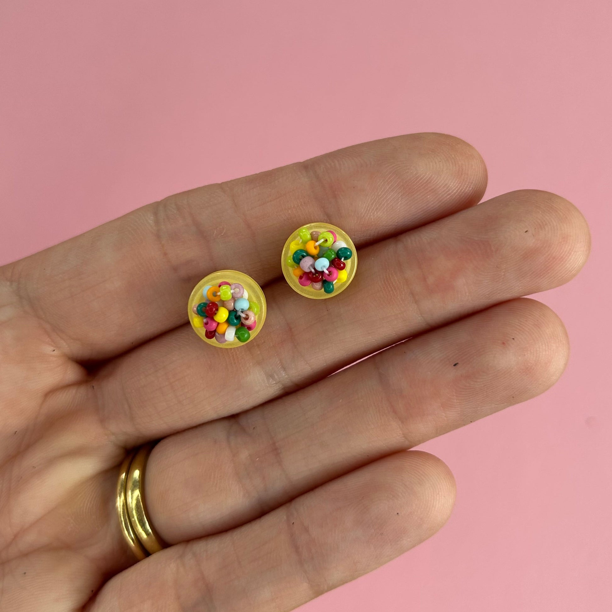 A close-up of a person's hand holding two small round yellow stud earrings filled with colorful confetti, set against a solid pink background.