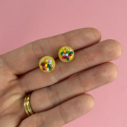 A close-up of a person's hand holding two small round yellow stud earrings filled with colorful confetti, set against a solid pink background.