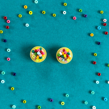 Two round stud earrings with colorful bead centers on a bright teal background, surrounded by scattered small multicolored beads.
