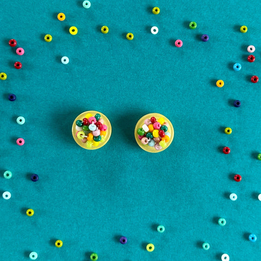 Two round stud earrings with colorful bead centers on a bright teal background, surrounded by scattered small multicolored beads.
